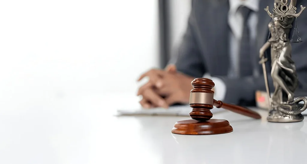 A gavel on a desk next to a Lady Justice statue, with a lawyer blurred in the background, representing legal action and justice for Lemon Law clients.