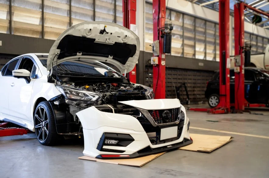 A white car with an open hood and missing front bumper in a garage, representing a vehicle under repair due to potential Lemon Law defects.